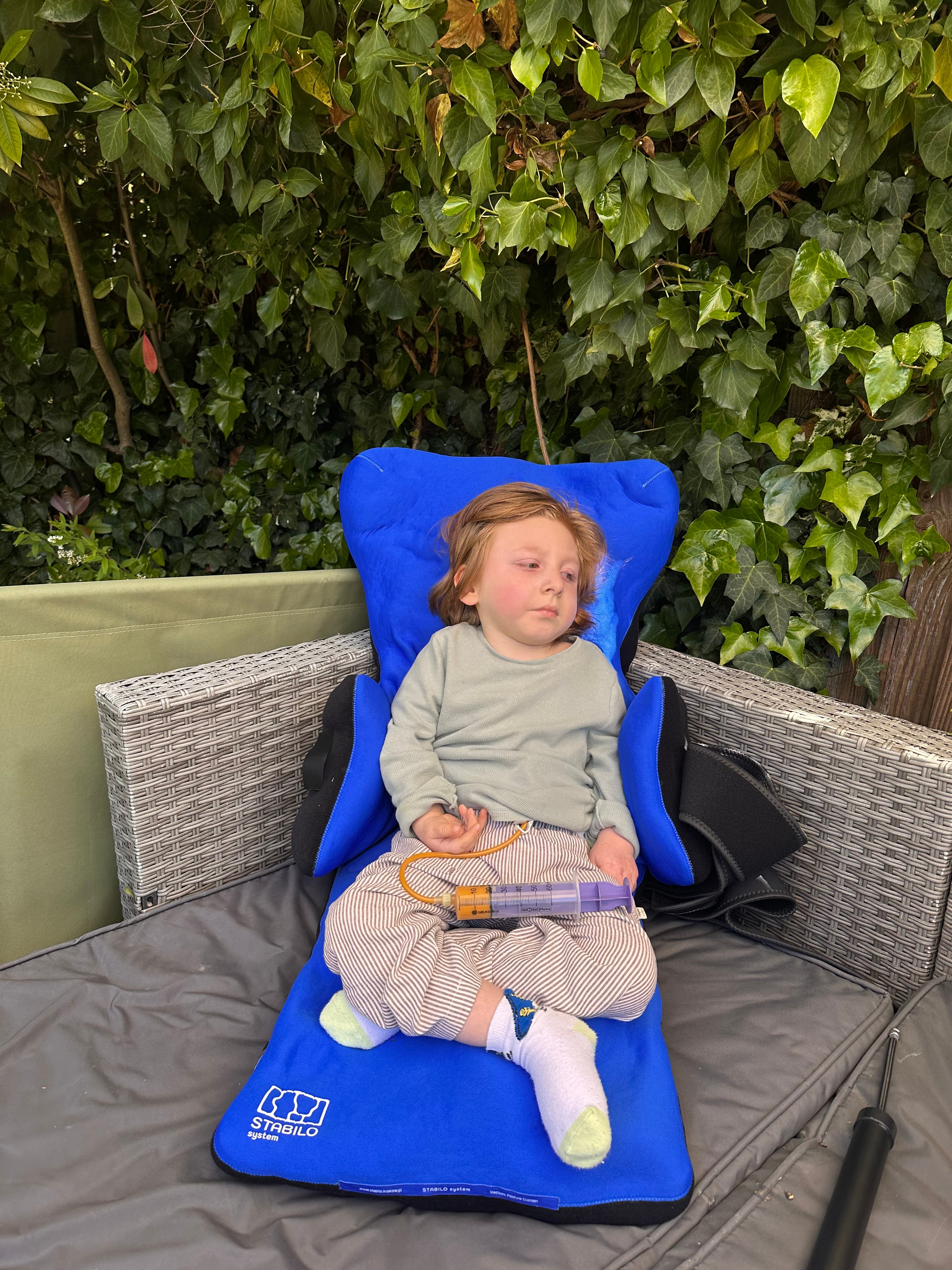 Young child seated in a blue supportive chair outdoors, with a large syringe resting on their lap and green ivy in the background.
