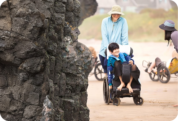 A caregiver pushing a smiling child in a beach wheelchair along a coastal path